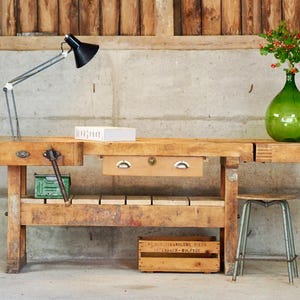 Könnte beinhalten: Eine Werkbank aus Holz mit einer mittigen Schublade mit zwei Metallgriffen, einem seitlichen Schraubstock mit Holzgriff und einem vorderen Schraubstock. Eine schwarze verstellbare Schreibtischlampe, ein weißes Buch und eine grüne Glasvase.