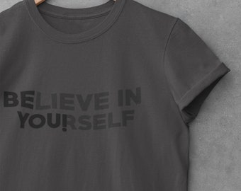 May include: A charcoal grey short-sleeve t-shirt featuring the text BELIEVE IN YOURSELF printed in a darker shade of grey across the chest. The shirt has a crew neckline and rolled sleeves, displayed on a wooden hanger.