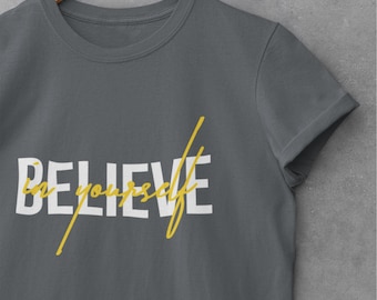 May include: A dark grey short-sleeved t-shirt featuring the word BELIEVE in large white capital letters. Overlaid across the white text is the phrase in yourself written in a yellow cursive script font. The shirt has a crew neckline.