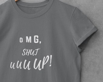 Puede incluir: Una camiseta gris oscuro de manga corta con texto impreso en blanco que dice "OMG, SHUT UUU UP!" en una mezcla de fuentes tipo bloque y manuscrita. La prenda tiene cuello redondo y se muestra en una percha de madera.