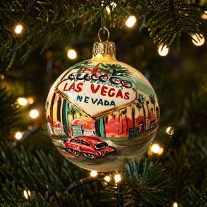 Könnte beinhalten: Eine runde Weihnachtskugel mit goldener Kappe, die an einem Weihnachtsbaum hängt. Die Kugel zeigt eine gemalte Szene von Las Vegas, Nevada, mit einem roten Auto und dem Text "Las Vegas Nevada". Der Hintergrund ist eine Mischung aus Grün, Gelb und Rot.
