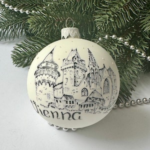 May include: White Christmas bauble with a black and white illustration of a cityscape and the word "Vienna" written below.