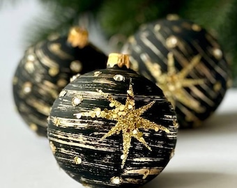 May include: Three black Christmas ornaments with gold glitter accents. The ornaments have a star design and are on a white background.