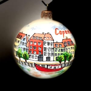 May include: A round Christmas ornament with a hand-painted scene of Copenhagen, Denmark. The ornament features colorful buildings, a red boat, and the word "Copenhagen" in red. The ornament is suspended against a black background.