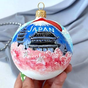 Puede incluir: Adorno navideño de cristal pintado a mano con un paisaje japonés. El adorno tiene un cielo azul con la palabra "JAPAN" y una montaña roja. La mitad inferior es rosa con flores de cerezo blancas.