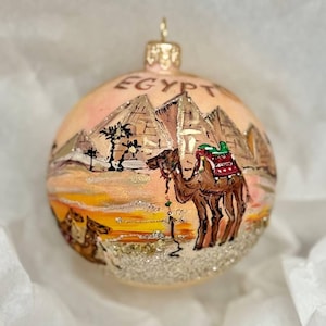 Op de afbeelding: Een ronde glazen kerstbal met een beige achtergrond en gouden glitter. De kerstbal heeft een geschilderd tafereel van de piramides van Egypte met een kameel en palmbomen. Het woord "EGYPT" staat in bruine letters op de kerstbal.
