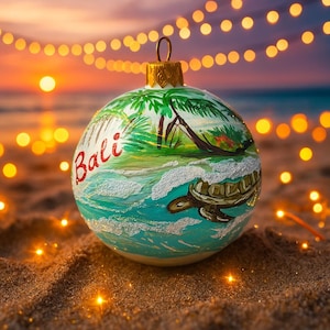 Peut inclure: Une boule de Noël ronde avec une scène de plage de Bali. L'ornement présente un paysage peint avec des palmiers, des vagues océaniques et une tortue de mer. Le mot "Bali" est écrit en script rouge. L'ornement est sur une surface sablonneuse.