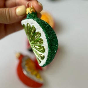 May include: A green and white glass Christmas ornament shaped like a lime wedge. The ornament is covered in glitter and has a gold top.