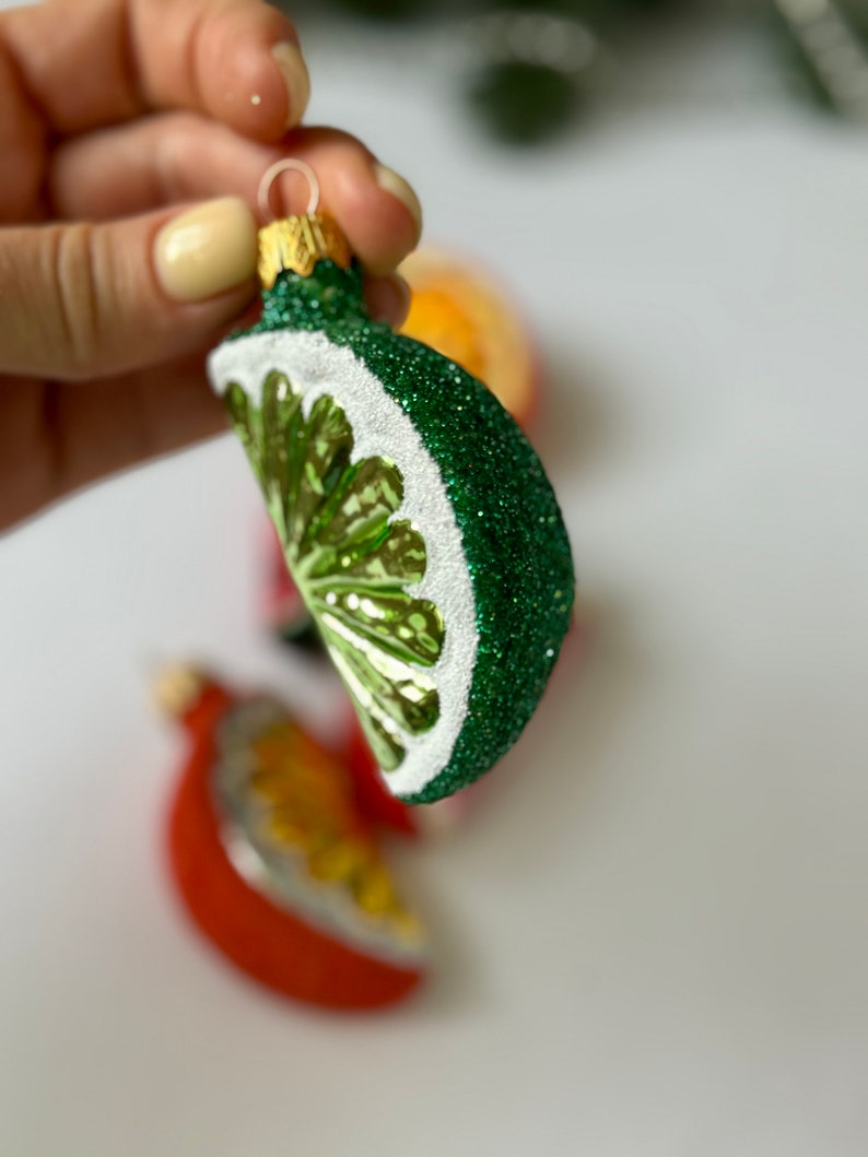 May include: A green and white glass Christmas ornament shaped like a lime wedge. The ornament is covered in glitter and has a gold top.