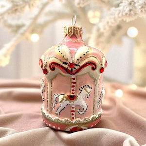 Pu&ograve; includere: Ornamento natalizio a forma di giostra rosa, con dettagli dorati e rossi. L'ornamento presenta cavalli bianchi e un design decorativo. La palla ha un anello di metallo dorato per appenderla. Una decorazione festiva.