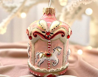 May include: Pink carousel Christmas ornament with gold and red accents. The ornament features white horses and a decorative design. The ornament has a gold metal loop for hanging. A festive holiday decoration.