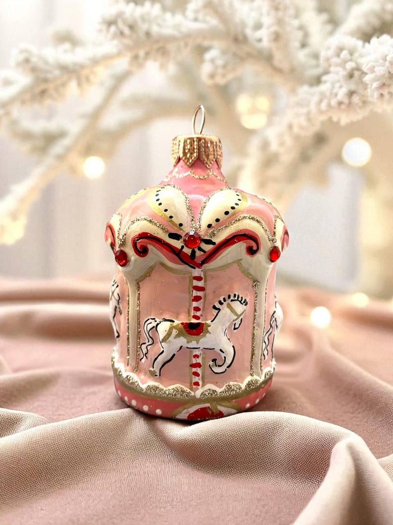 Pu&ograve; includere: Ornamento natalizio a forma di giostra rosa, con dettagli dorati e rossi. L'ornamento presenta cavalli bianchi e un design decorativo. La palla ha un anello di metallo dorato per appenderla. Una decorazione festiva.