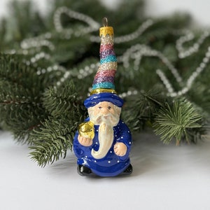 May include: A blue glass Christmas ornament depicting a wizard with a white beard and a sparkly pink, blue, and white hat. He is holding a gold bell in his right hand.