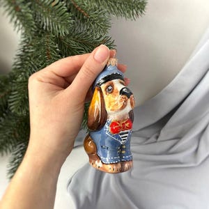 Peut inclure: Une boule de Noël en verre en forme de chien portant une tenue de marin bleue avec un nœud papillon rouge. Le chien a le pelage brun et la poitrine blanche.