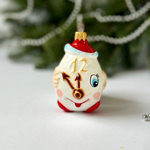 Puede incluir: Adorno navideño caprichoso con forma de reloj, con una combinación de colores rojo y blanco. La esfera del reloj tiene una cara sonriente, manecillas que apuntan a las 12 y mejillas rosadas. El adorno está suspendido sobre un fondo verde borroso.