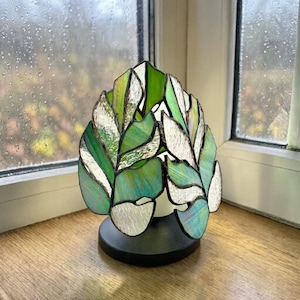 Op de afbeelding: Een gebrandschilderde lamp in de vorm van bladeren, met tinten groen en wit. De lamp staat op een zwarte voet en staat op een houten oppervlak bij een raam.