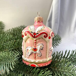 Pu&ograve; includere: Ornamento natalizio a forma di giostra rosa e bianco con dettagli dorati e rossi. L'ornamento presenta un design a giostra con cavalli bianchi, su uno sfondo di rami di pino.