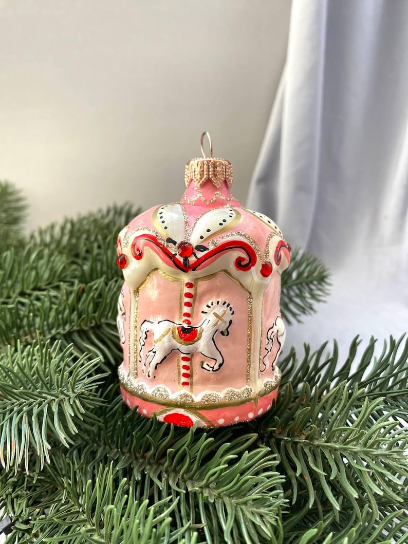 Pu&ograve; includere: Ornamento natalizio a forma di giostra rosa e bianco con dettagli dorati e rossi. L'ornamento presenta un design a giostra con cavalli bianchi, su uno sfondo di rami di pino.
