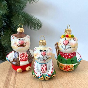 Op de afbeelding: Drie handgeschilderde glazen kerstversieringen in de vorm van katten die traditionele Oekraïense kleding dragen. De versieringen zijn wit met rode, groene en bruine accenten.