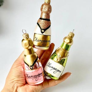 Puede incluir: Tres adornos navideños de vidrio con forma de botellas de champán. Cada botella es de un color diferente: rosa, verde y dorado. Las botellas tienen detalles dorados y la palabra "Champagne" escrita en ellas.