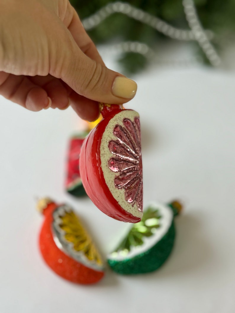 May include: Four glass ornaments shaped like fruit slices. The ornaments are red, green, and yellow. The ornaments are decorated with glitter.