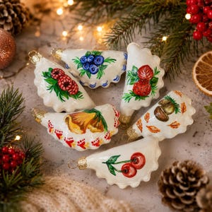 May include: A collection of white, scalloped-edged Christmas ornaments, each hand-painted with a different fruit or botanical design. The ornaments feature cherries, raspberries, blueberries, strawberries, and a yellow flower. Gold-toned caps and hangers complete the festive look.