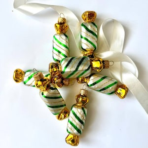 May include: Six candy cane-striped Christmas ornaments in the shape of wrapped candy. The ornaments are white with green and gold stripes and have gold accents.