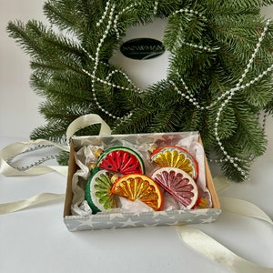 May include: A set of five glass Christmas ornaments shaped like fruit slices. The ornaments are red, orange, yellow, and green. They are in a white box with a silver star pattern. The box is sitting on a white surface.