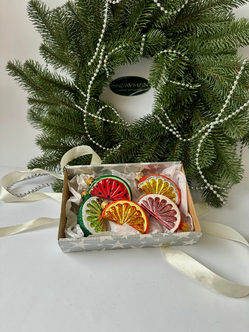 May include: A set of five glass Christmas ornaments shaped like fruit slices. The ornaments are red, orange, yellow, and green. They are in a white box with a silver star pattern. The box is sitting on a white surface.