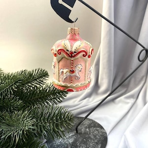 Pu&ograve; includere: Un ornamento natalizio a forma di giostra rosa e bianca con dettagli dorati e dettagli di cavalli bianchi. L'ornamento &egrave; sospeso da un supporto in metallo nero con base circolare, accanto a un rametto di sempreverde.