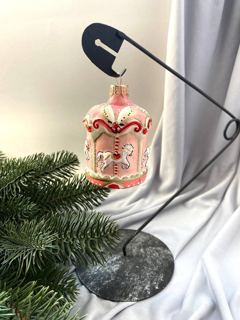 Pu&ograve; includere: Un ornamento natalizio a forma di giostra rosa e bianca con dettagli dorati e dettagli di cavalli bianchi. L'ornamento &egrave; sospeso da un supporto in metallo nero con base circolare, accanto a un rametto di sempreverde.