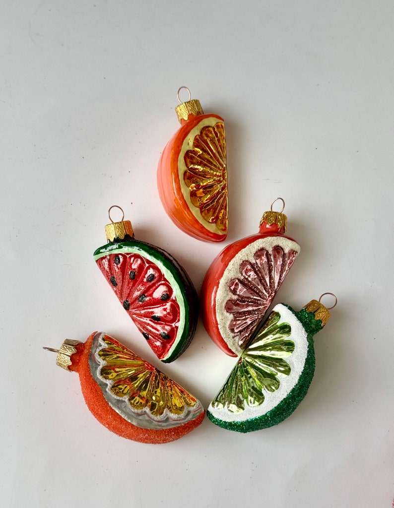 May include: Five glass Christmas ornaments shaped like fruit slices. The ornaments are red, green, and orange with gold accents. The ornaments are a watermelon slice, a lime slice, a grapefruit slice, and two orange slices.