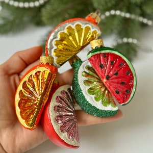May include: A set of five glass Christmas ornaments shaped like fruit slices. The ornaments are red, green, yellow, and orange. They are decorated with glitter and have a white background.