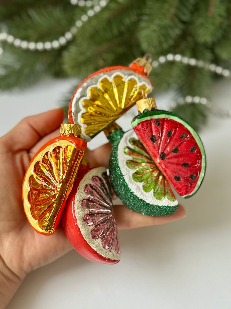 May include: A set of five glass Christmas ornaments shaped like fruit slices. The ornaments are red, green, yellow, and orange. They are decorated with glitter and have a white background.