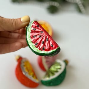May include: A set of four glass ornaments shaped like fruit slices. The ornaments are red, green, and yellow and feature a watermelon, lime, and orange slice.