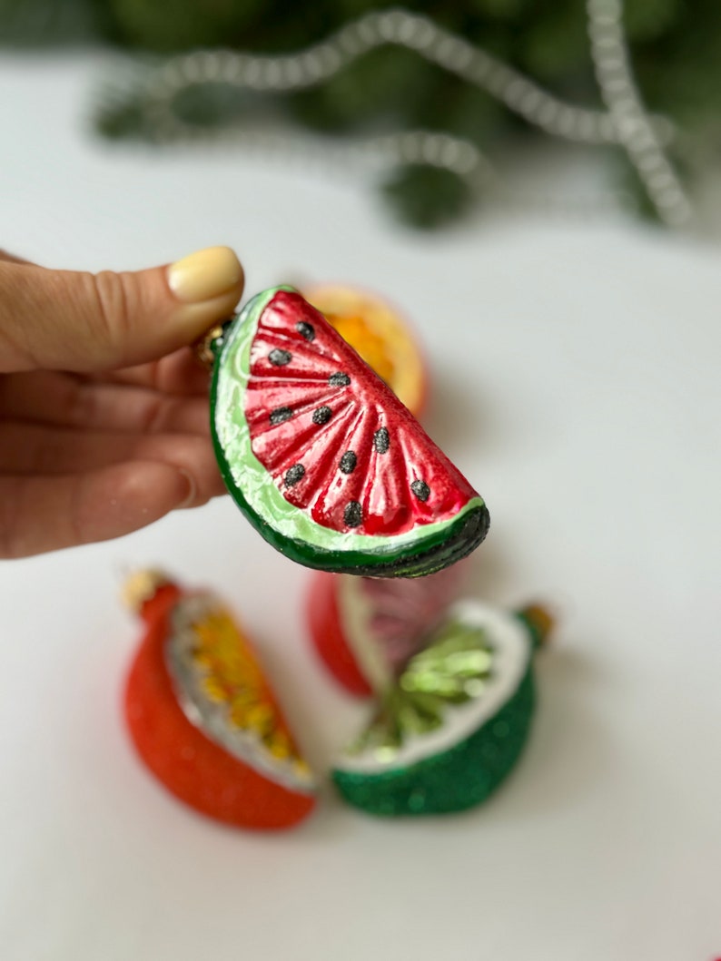 May include: A set of four glass ornaments shaped like fruit slices. The ornaments are red, green, and yellow and feature a watermelon, lime, and orange slice.