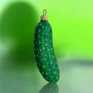 May include: A green glass Christmas ornament shaped like a pickle. The ornament has a gold-colored metal cap and a loop for hanging. The pickle has a textured surface with small, light-colored bumps.
