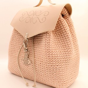 Puede incluir: Una mochila de crochet rosa claro con una solapa de cuero y un asa de cuero. La solapa tiene un dise&ntilde;o decorativo de c&iacute;rculos y el texto "Anem Veloni" est&aacute; estampado en la solapa.