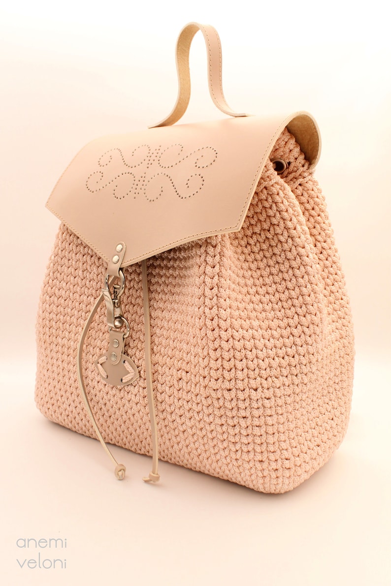 Puede incluir: Una mochila de crochet rosa claro con una solapa de cuero y un asa de cuero. La solapa tiene un dise&ntilde;o decorativo de c&iacute;rculos y el texto "Anem Veloni" est&aacute; estampado en la solapa.