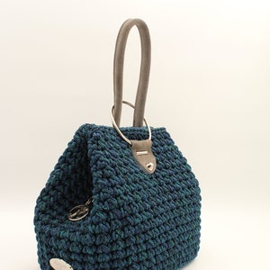 May include: A blue and green crocheted handbag with a gray suede handle and a silver ring top closure. The bag has a silver metal tag with the text "anemi veloni" on it.
