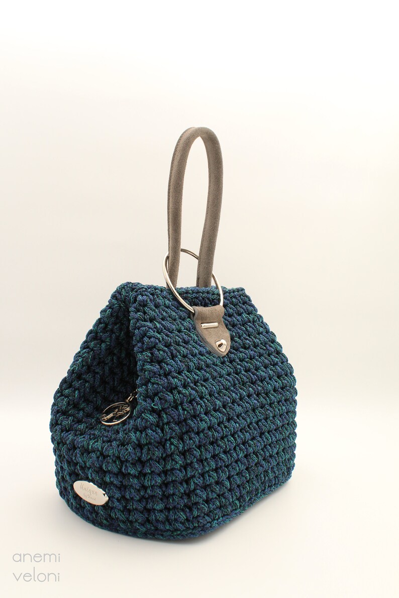 May include: A blue and green crocheted handbag with a gray suede handle and a silver ring top closure. The bag has a silver metal tag with the text "anemi veloni" on it.