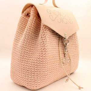 Puede incluir: Una mochila de crochet rosa claro con una solapa de cuero y un cierre met&aacute;lico decorativo. La solapa tiene un dise&ntilde;o de coraz&oacute;n con un patr&oacute;n circular.
