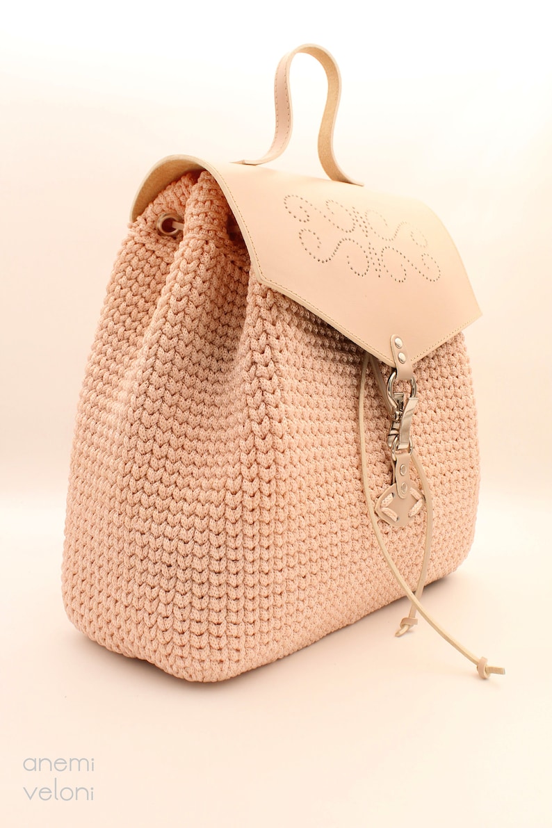 Puede incluir: Una mochila de crochet rosa claro con una solapa de cuero y un cierre met&aacute;lico decorativo. La solapa tiene un dise&ntilde;o de coraz&oacute;n con un patr&oacute;n circular.