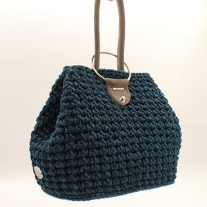 May include: A dark blue and green crocheted handbag with a silver ring handle and a gray suede strap.