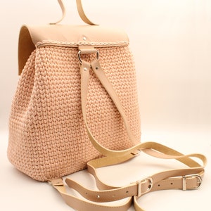 Puede incluir: Una mochila de crochet rosa claro con correas de cuero y una solapa de cuero. La mochila tiene un asa superior y correas ajustables.