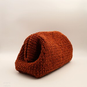 May include: A rust-coloured crocheted bag with a textured surface. The bag is folded in half and is sitting on a white surface.