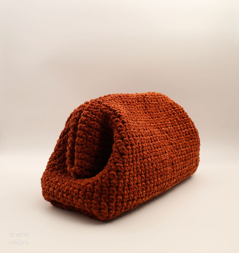 May include: A rust-coloured crocheted bag with a textured surface. The bag is folded in half and is sitting on a white surface.