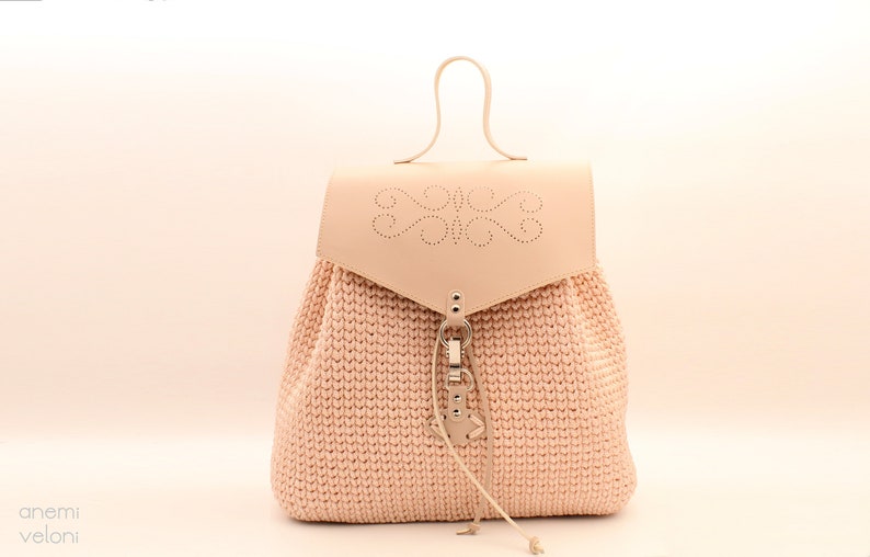Puede incluir: Una mochila de crochet rosa claro con una solapa de cuero y un cierre met&aacute;lico decorativo. La solapa tiene un dise&ntilde;o recortado.
