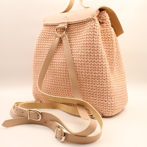 Puede incluir: Una mochila de crochet rosa claro con correas y hebillas de cuero. La mochila tiene un cierre de solapa y un asa superior.