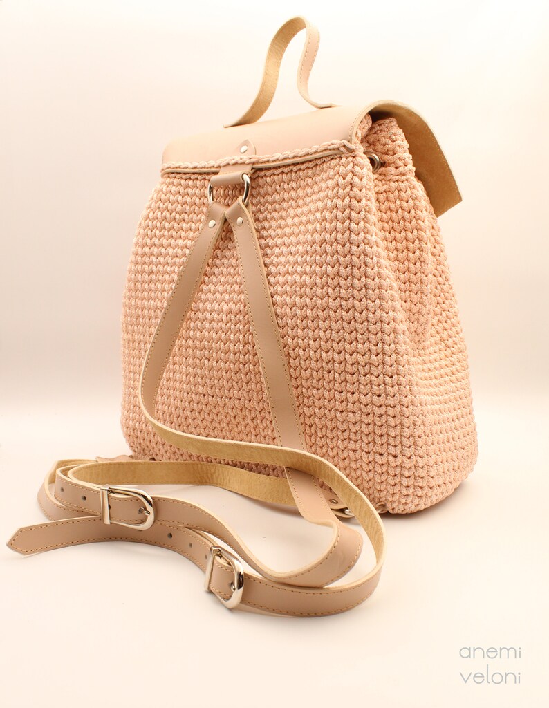 Puede incluir: Una mochila de crochet rosa claro con correas y hebillas de cuero. La mochila tiene un cierre de solapa y un asa superior.
